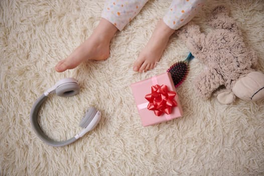 Top view child's feet on a carpet near wireless headphones, hairbrush, gift box and plush toy