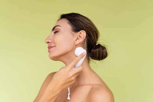 Beauty portrait of young topless woman with bare shoulders on green background with perfect skin and natural makeup holds face cleansing brush