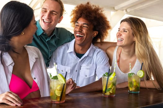 Group of happy multiracial friends laughing and having fun drinking mojito cocktails at a beach bar.