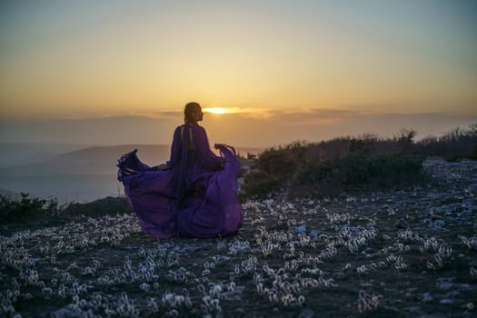 Sunset purple dress woman mountains. Rise of the mystic. sunset over the clouds with a girl in a long purple dress. In the meadow there is a grass dream with purple flowers.