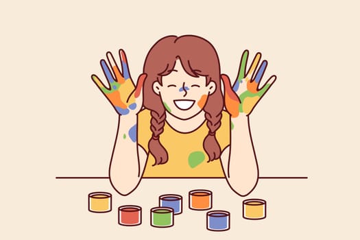 Positive girl with face and palms smeared with multi-colored paint smiles, wanting to become artist