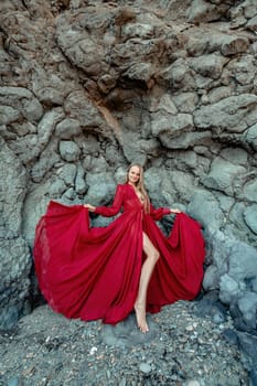 Red dress rocks woman. A blonde with flowing hair in a long flowing red dress stands near a rock of volcanic origin. Travel concept, photo session at sea