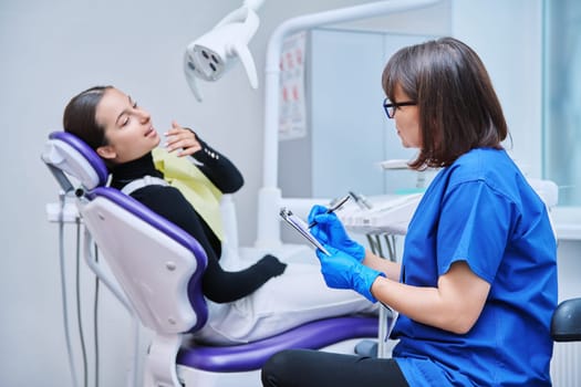 Dentist and teenage girl patient in dental clinic, doctor nurse writing on clipboard consulting, teen female talking about toothache disturbing dental problem. Dentistry, treatment, dental health care