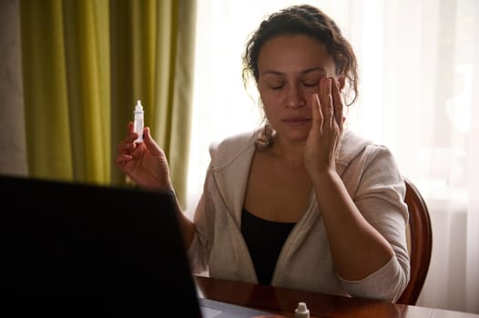 Multiethnic woman sitting at table with laptop and using medicinal eye drops to relieve dry eyes and digital eye strain