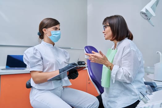 Dentist and woman patient in dental clinic, doctor nurse writing on clipboard consulting, middle aged female talking about disturbing dental problem. Dentistry, treatment, dental health care concept