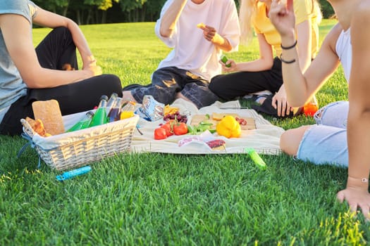 Close-up food and drink for picnic on green grass. Group of young people resting in nature, basket with bread, cheese fruits vegetables snacks juices on tablecloth. Youth, leisure, lifestyle, fun,