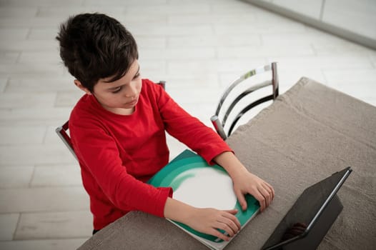 Overhead view of a school child doing homework with digital tablet at home. Kid using gadgets to study. Education and distance learning for kids. Homeschooling during quarantine. Online courses