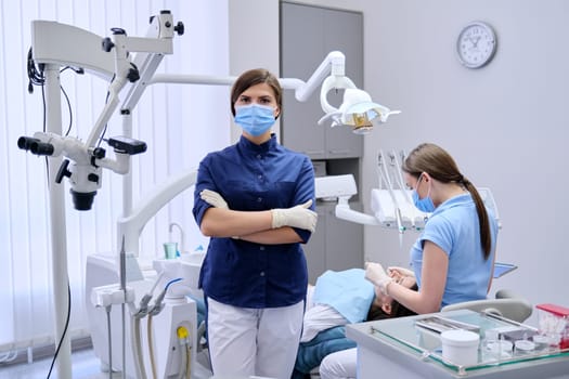 Portrait of young female doctor dentist in protective medical face mask with arms crossed in dental office, treating woman patient in chair background. Medicine, dentistry and health care concept