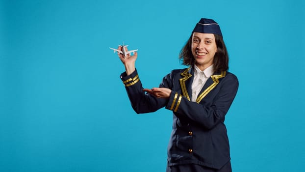 Professional air hostess presenting miniature toy plane, feeling happy about flying occupation. Young woman working as stewardess in uniform showing small artificial airplane on camera.