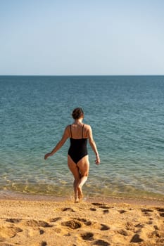 Woman sea swimsuit sand. A girl in full growth stands with her back and enters the sea in a black swimsuit. Alone on the beach on a sunny day.