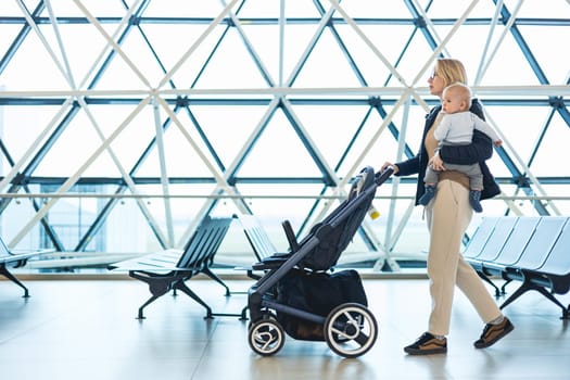 Mother carying his infant baby boy child, pushing stroller at airport departure terminal moving to boarding gates to board an airplane. Family travel with baby concept.