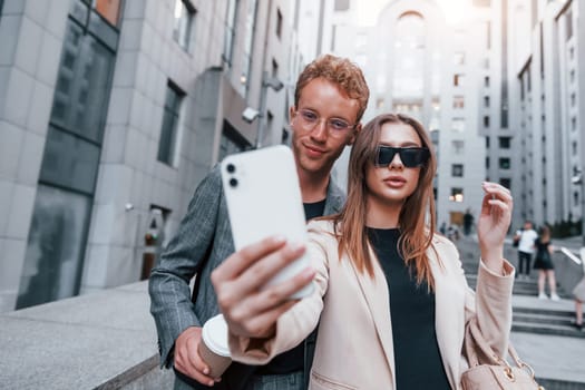 Makes selfie. Woman and man in the town at daytime. Well dressed people