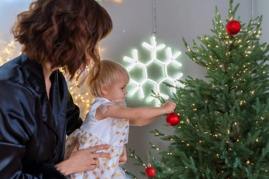 A mother with a 2-year-old daughter decorates the Christmas tree. Mom in a black suit, a girl in a white dress, her daughter hangs a red ball on the Christmas tree. Merry Christmas and New Year concept