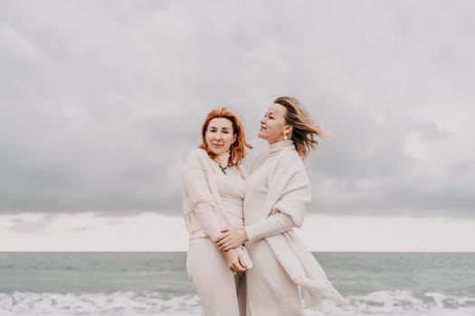Women sea walk friendship spring. Two girlfriends, redhead and blonde, middle-aged walk along the sandy beach of the sea, dressed in white clothes. Against the backdrop of a cloudy sky and the winter sea. Weekend concept.