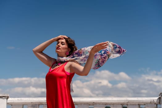 A woman in a red dress against a blue sky and white clouds with a developing scarf around her neck. She turned her face to the wind and posed for the camera.