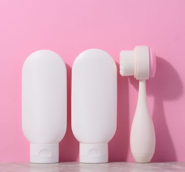 White plastic tubes with cream and a massaging brush for facial cleansing on a pink background, items for cosmetic procedures
