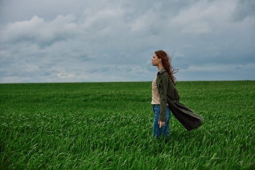 a beautiful woman stands in a green field in a raincoat and looks into the distance. Strong wind, flying hair, harmony with nature