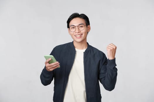 Excited young Asian man using smartphone and making winner gesture isolated 
