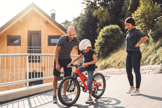 Dad teaching her young daughter how to ride a bike, while mom stands by looking at them