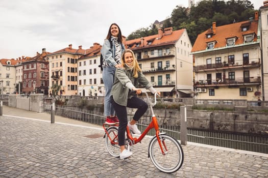 Couple of best friends riding a bike on the city streets, one of the women standing on the trunk of the bike