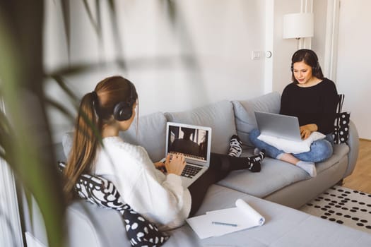 Two young women students studying on their laptops on the couch in their apartment each with their headphones on