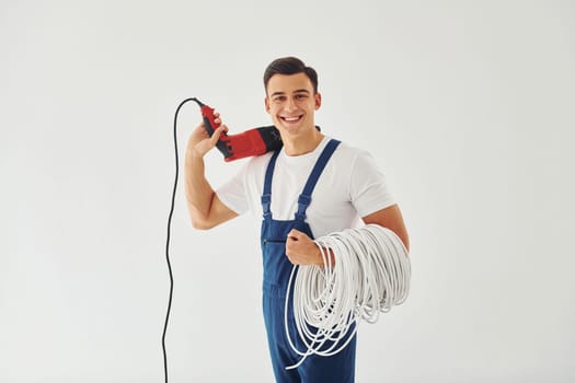 With drill and cables in hands. Male worker in blue uniform standing inside of studio against white background