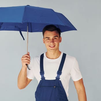 Holds umbrella by hand. Male worker in blue uniform standing inside of studio against white background
