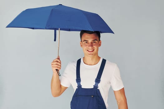 Holds umbrella by hand. Male worker in blue uniform standing inside of studio against white background
