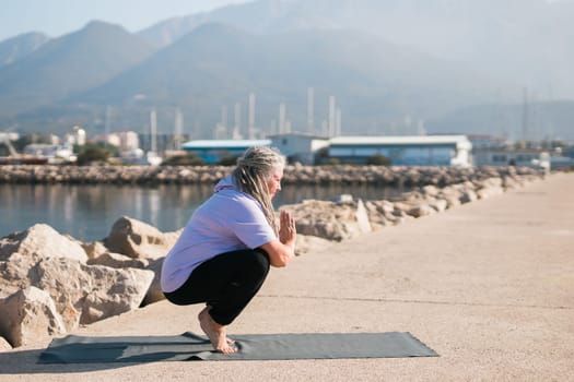 Mature woman with dreadlocks working out doing yoga exercises on sea beach copy space - wellness well-being and active elderly age concept