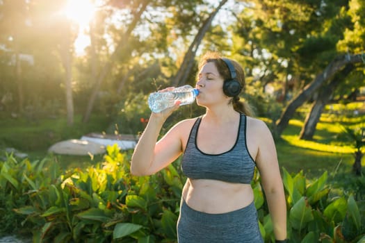 Overweight woman drinking water after jogging in the park. Portrait of young plus-size thirsty woman with a bottle of water outdoors copy space. Sports healthcare and weight loosing fitness and well-being concept