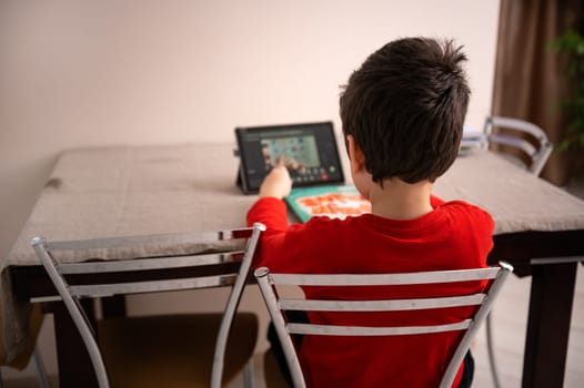 Rear view of a school child doing homework, sitting at table and watching online lesson on a digital tablet