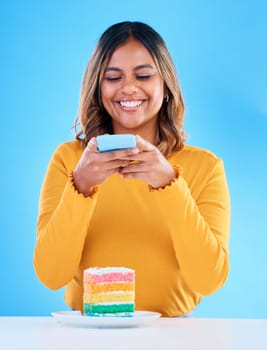 Woman, cake and studio with a phone for social media picture with a smile and excited to eat. Happy female person on blue background with rainbow color dessert for birthday or influencer celebration.