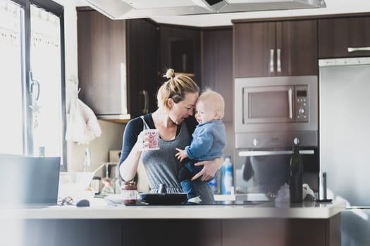 Happy mother holding her little infant baby boy while drinking morning coffee and making pancakes for breakfast in domestic kitchen. Family lifestyle, domestic life concept.