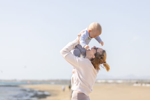 Mother enjoying summer vacations holding, playing and lifting his infant baby boy son high in the air on sandy beach on Lanzarote island, Spain. Family travel and vacations concept.