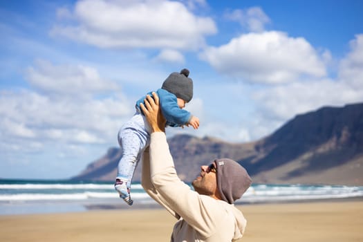 Father enjoying pure nature holding and playing with his infant baby boy son in on windy sandy beach of Famara, Lanzarote island, Spain. Family travel and parenting concept.