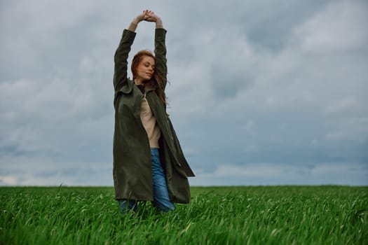 a woman in a long coat stands in a field against a cloudy sky with her back to the wind with her hands raised behind her head