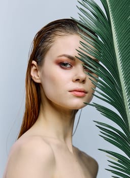 a close beauty portrait of a beautiful woman standing on a light background holding a palm leaf near her face, looking into the camera. Vertical photo without retouching of problem skin. High quality photo