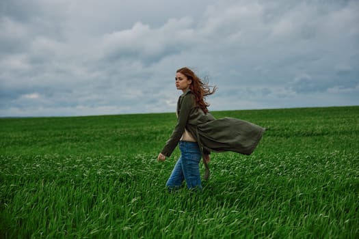 a beautiful woman stands in a green field in a raincoat and looks into the distance. Strong wind, flying hair, harmony with nature