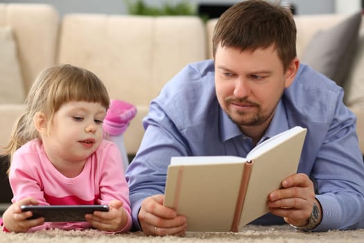 Dad reads a book to daughter holding smartphone lying on floor