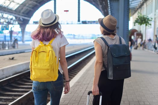 Mother and daughter teenager with backpacks suitcase walking in train station. Happy parent and child hiking together, women in hats, summer family vacation, back view