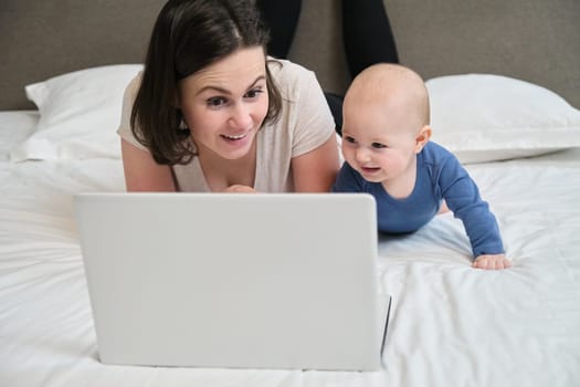 Woman with baby lying at home in bed looking with interest at laptop