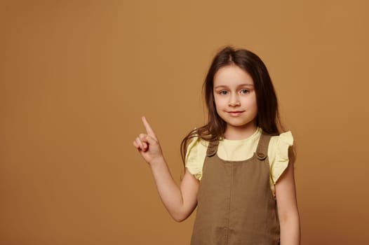 Happy preschooler child girl smiles looking at camera and points forefinger at copy space on isolated beige background