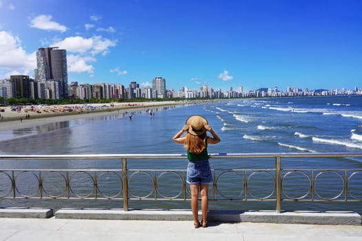 Full length of tourist girl holding hat enjoying view of skyscrapers skyline and waving ocean in Santos, Sao Paulo, Brazil