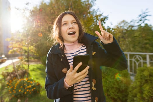 Young smiling teen girl shows index finger up, attention idea eureka, outdoor background, golden hour