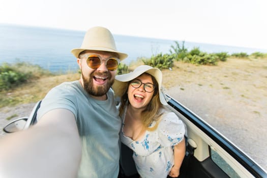 Romantic couple making selfie on smartphone camera in rental cabrio car on ocean or sea beach enjoying summer vacation together and taking picture on cellular resting near sea on weekends