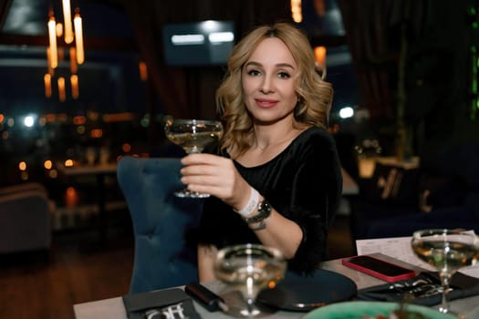 woman in a restaurant with champagne in her hand. She is sitting at a table against the backdrop of a dark restaurant hall with lamps.