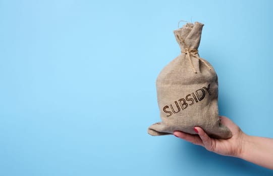 A female hand holds a burlap sack with the inscription subsidy on a blue background, concept of increasing government subsidies to the population