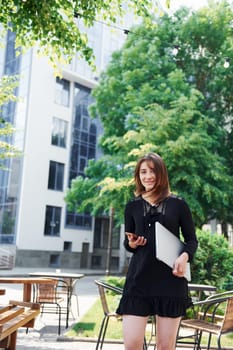 Holding laptop and phone. Happy young girl in black skirt outdoors in the city near green trees and against business building
