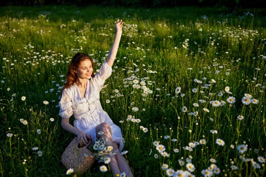 a beautiful woman in a light dress sits in a field of daisies with her hands raised above her head in the rays of the setting sun