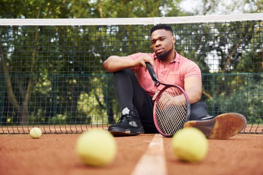 Sits near net and taking a break. African american man in pink shirt sits with tennis racket on the court outdoors